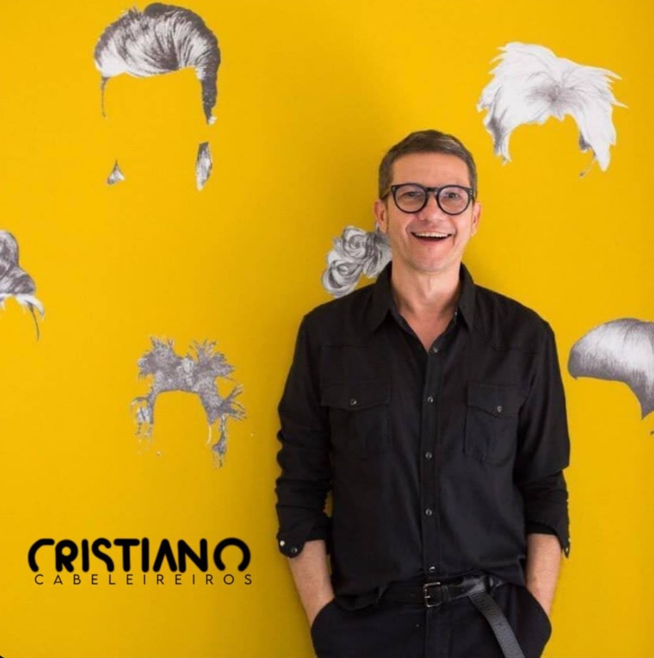 Retrato do Cristiano diante de uma parede amarela com o logo do salão