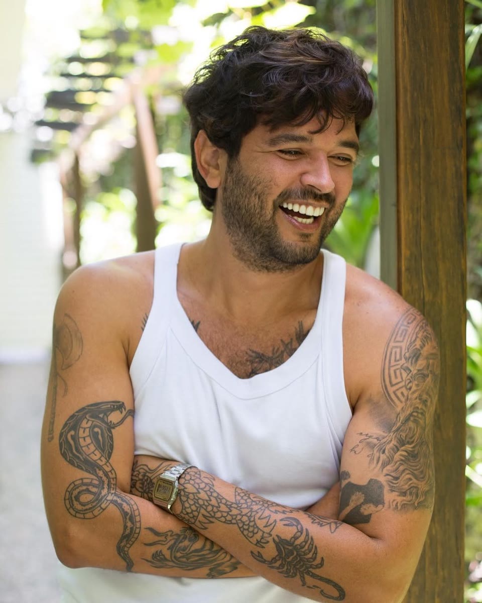 Alan rindo no jardim, braços cruzados, com cabelo cacheado e barba